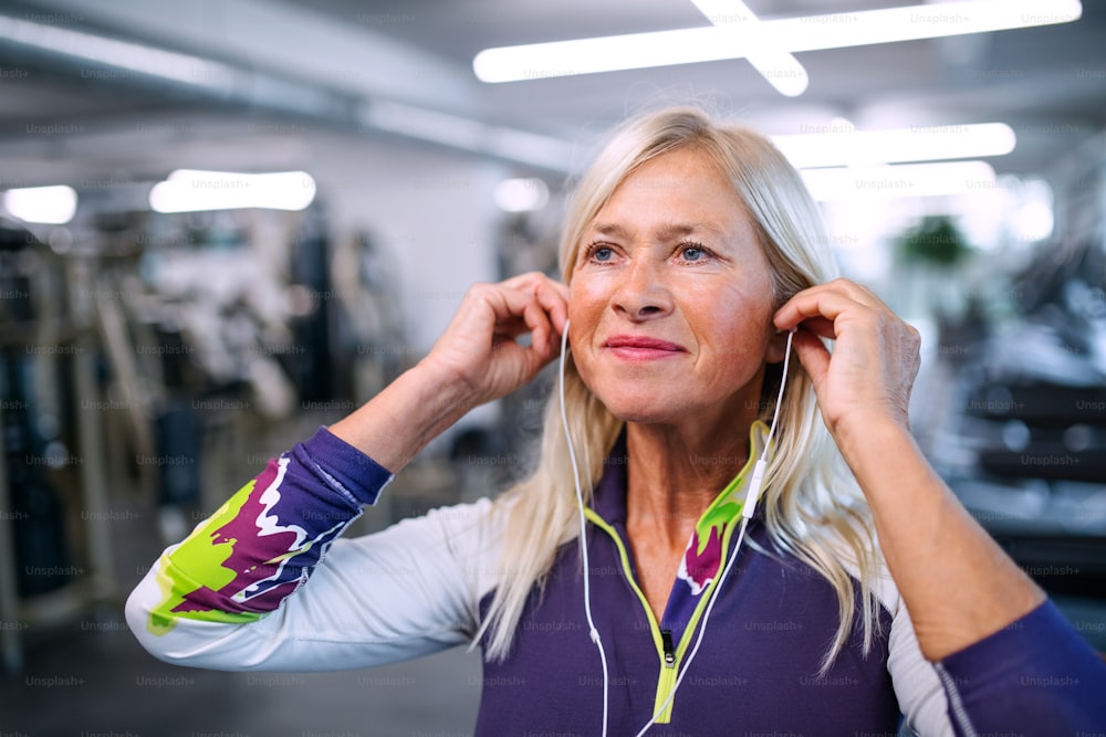 A front view of senior woman with earphones in gym resting after doing exercise.