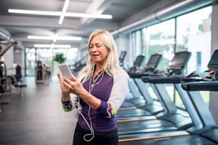 A front view of senior woman with earphones and smartphone in gym resting after doing exercise.