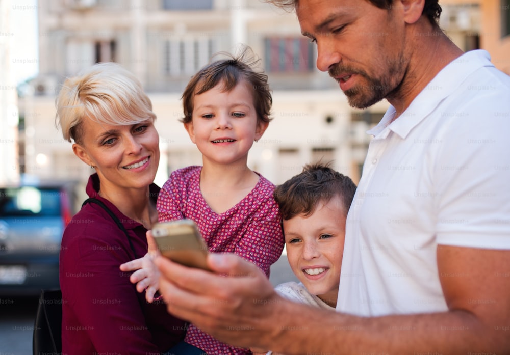 Una giovane famiglia con due bambini piccoli in piedi all'aperto in città, usando lo smartphone.