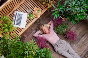 A top view of senior woman with laptop lying outdoors on terrace, resting.