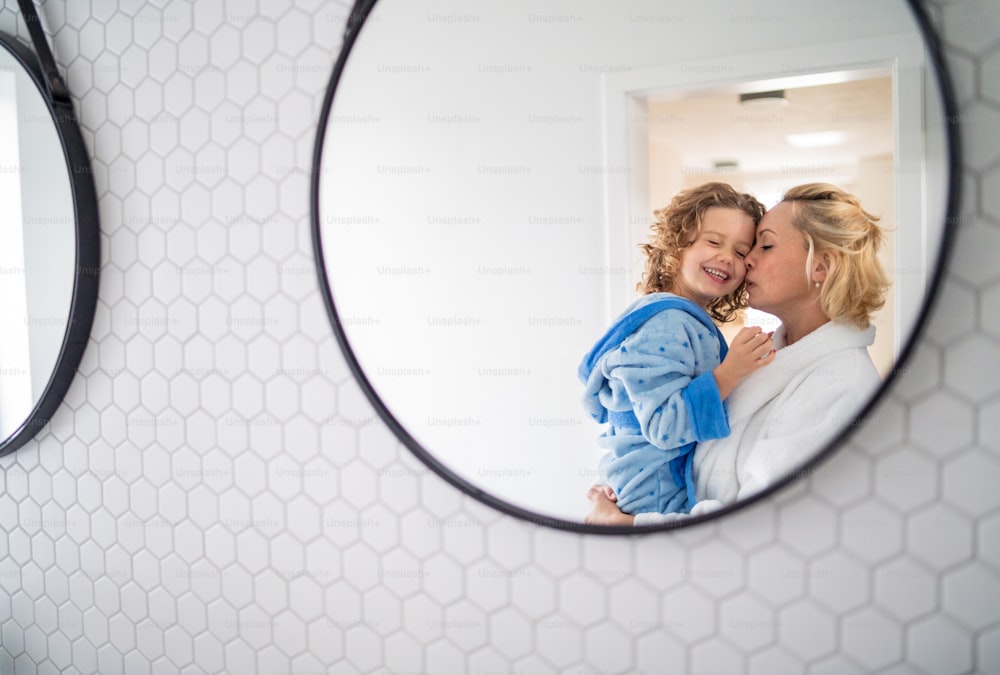 A reflection of mother and small daughter in mirror in bathroom indoors at home.