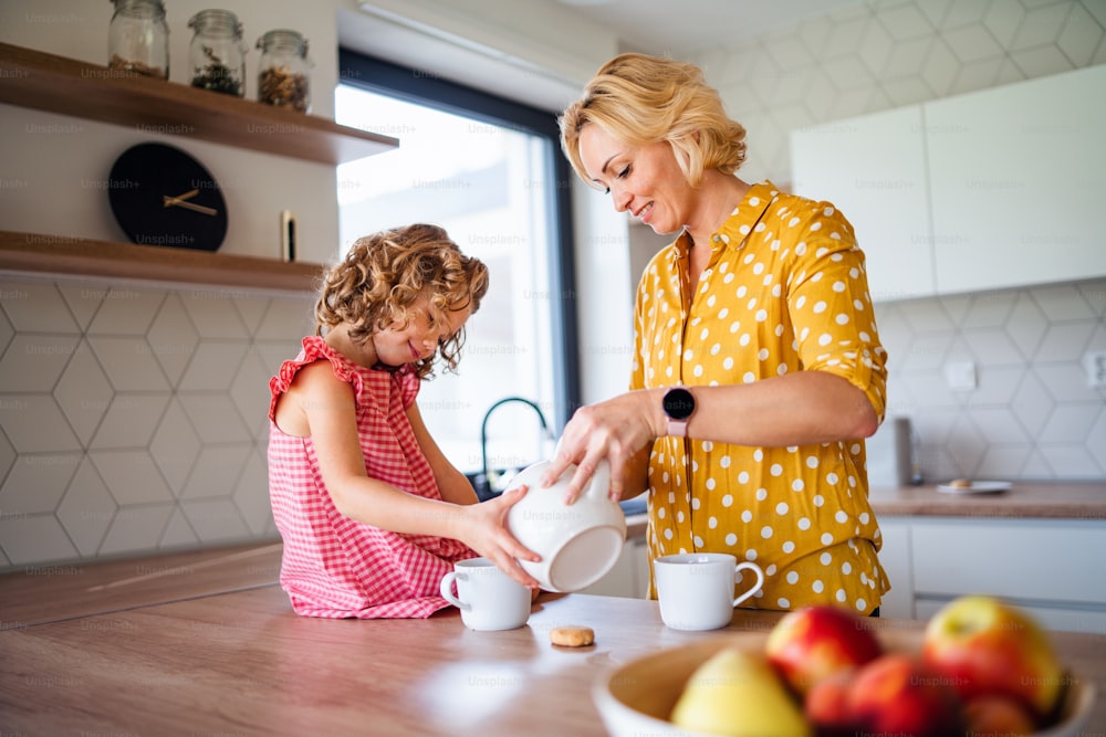 Une petite fille mignonne heureuse avec sa mère à l’intérieur dans la cuisine à la maison, buvant du thé.