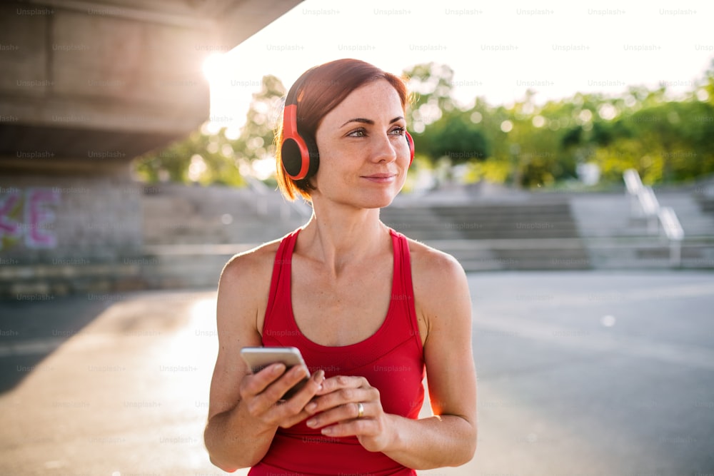 Young woman runner with headphones in city, using smartphone when resting.