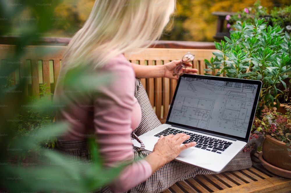 Une femme architecte chevronnée avec un ordinateur portable, assise à l’extérieur sur la terrasse, en train de travailler.