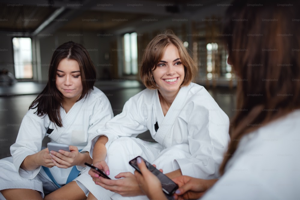 Un groupe de jeunes femmes karatés avec des smartphones à l’intérieur d’un gymnase, se reposant.