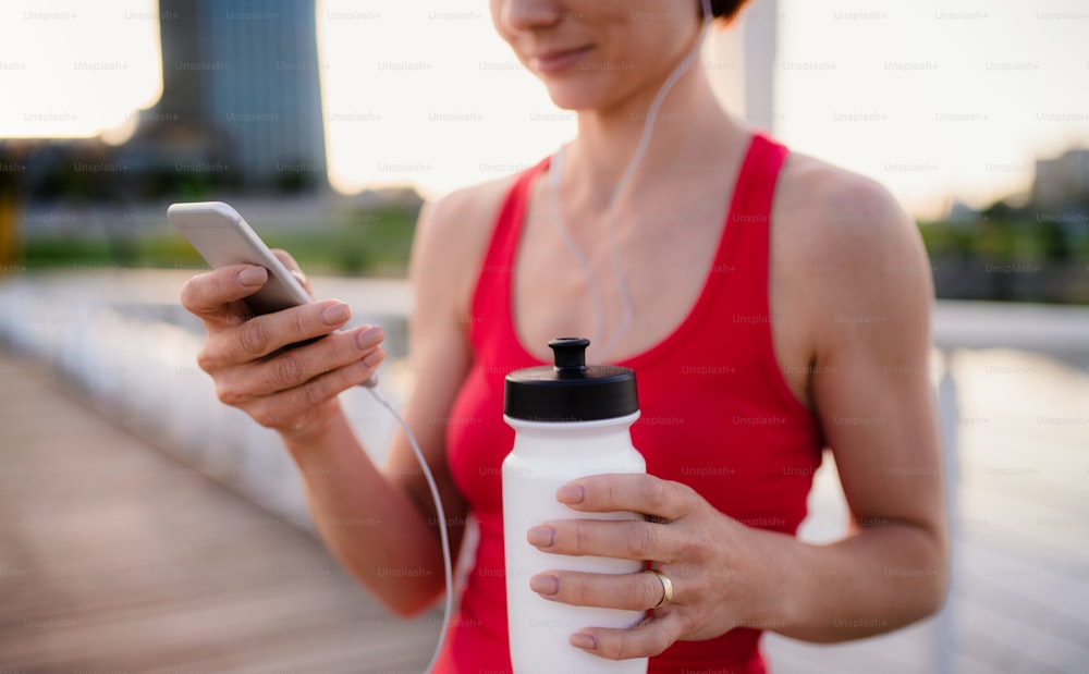 Midsection of young woman runner with earphones in city, using smartphone when resting.