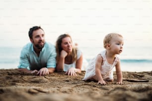 A family with a toddler girl lying on sand beach on summer holiday, playing.