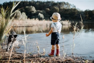 A rear view of small toddler boy with a hat and a dog standing by a lake. Copy space.