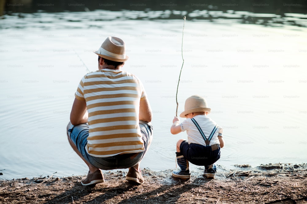 강이나 호숫가에서 야외에서 낚시를 하는 작은 유아 아들과 함께 있는 성숙한 아버지의 뒷모습.