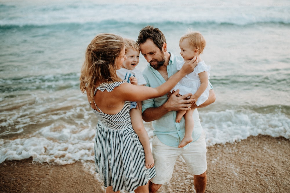 Una familia joven feliz con dos niños pequeños de pie en la playa durante las vacaciones de verano.