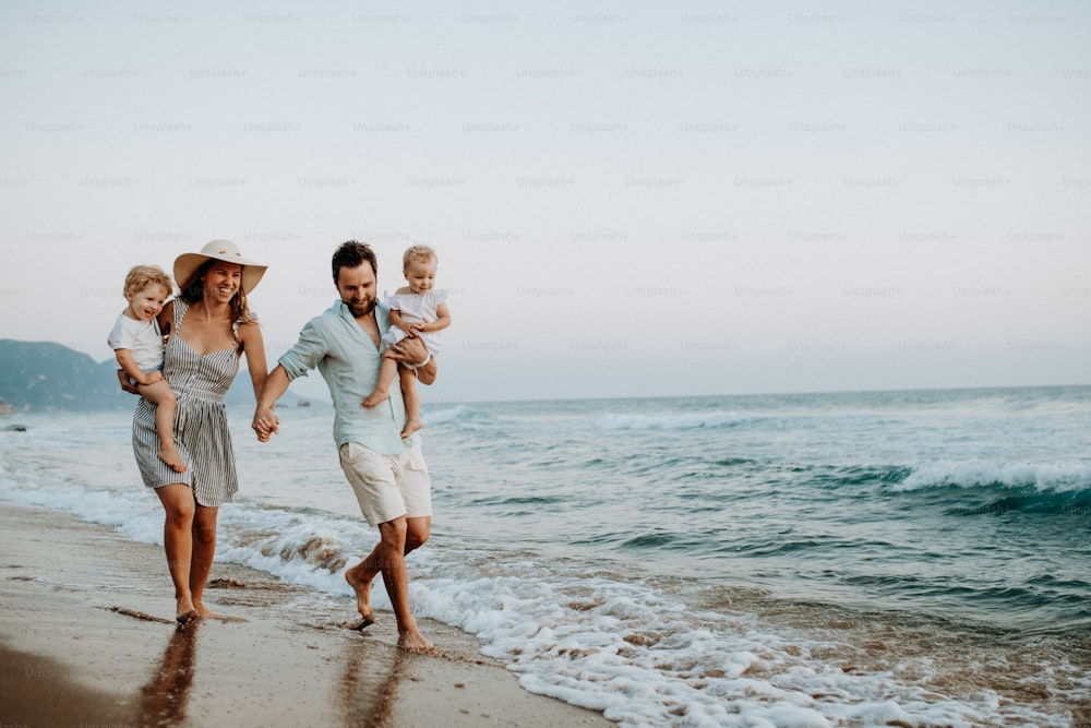 Uma família com duas crianças pequenas andando na praia nas férias de verão ao pôr do sol. Um pai e uma mãe carregando filho e filha nos braços.