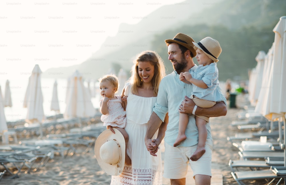 Eine junge Familie mit zwei Kleinkindern, die in den Sommerferien Händchen haltend am Strand spazieren gehen.