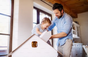 Mature father with small daughter playing with cardboard house indoors at home.