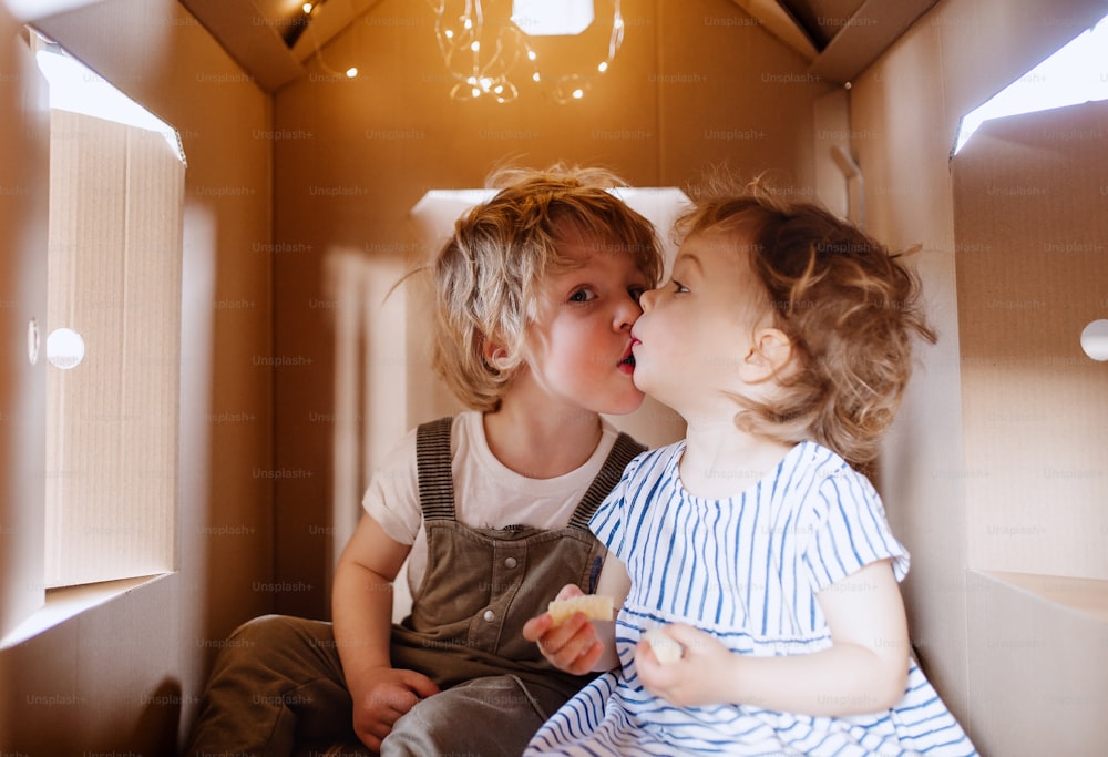 Two happy toddler children playing indoors in cardboard house at home, kissing.