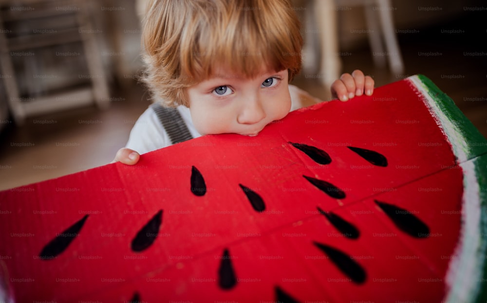 A close-up of toddler boy playing with large toy fruit indoors at home, eating fruit concept.