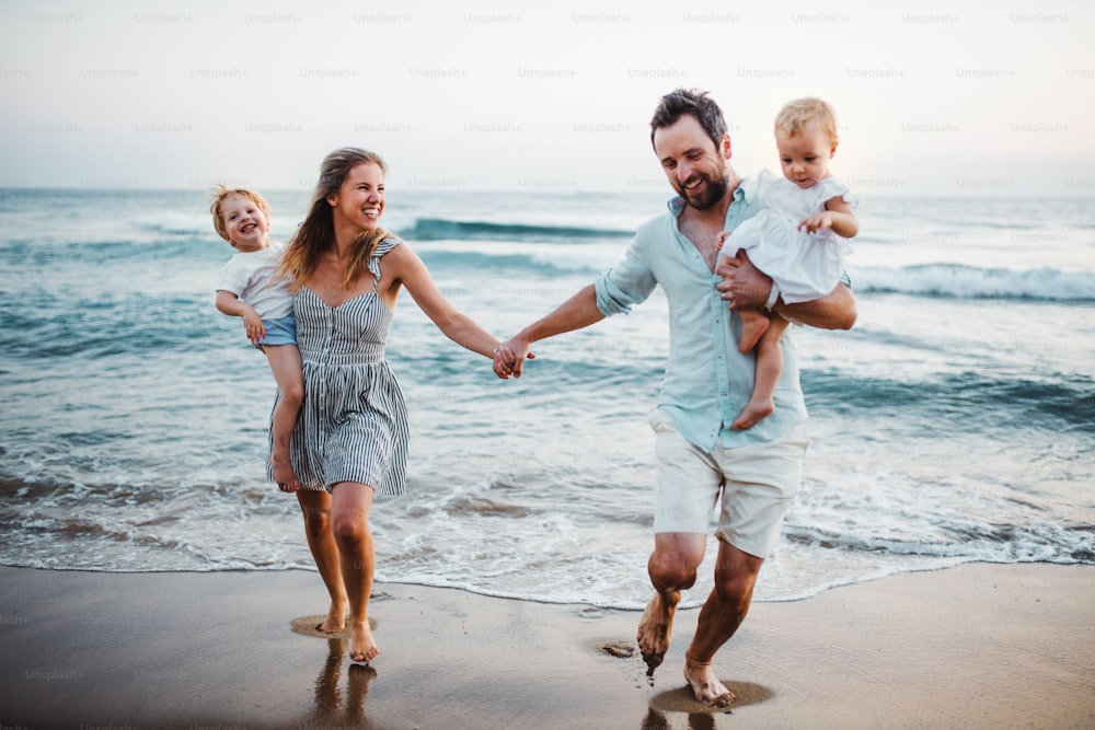 여름 휴가에 해변을 걷고 있는 두 명의 유아 자녀가 있는 젊은 가족이 손을 잡고 있습니다.