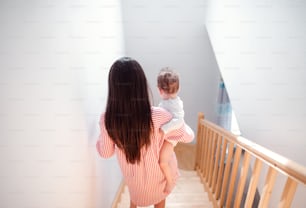 Vista trasera de una joven madre bajando las escaleras en el interior de la casa, cargando a su pequeño hijo pequeño.