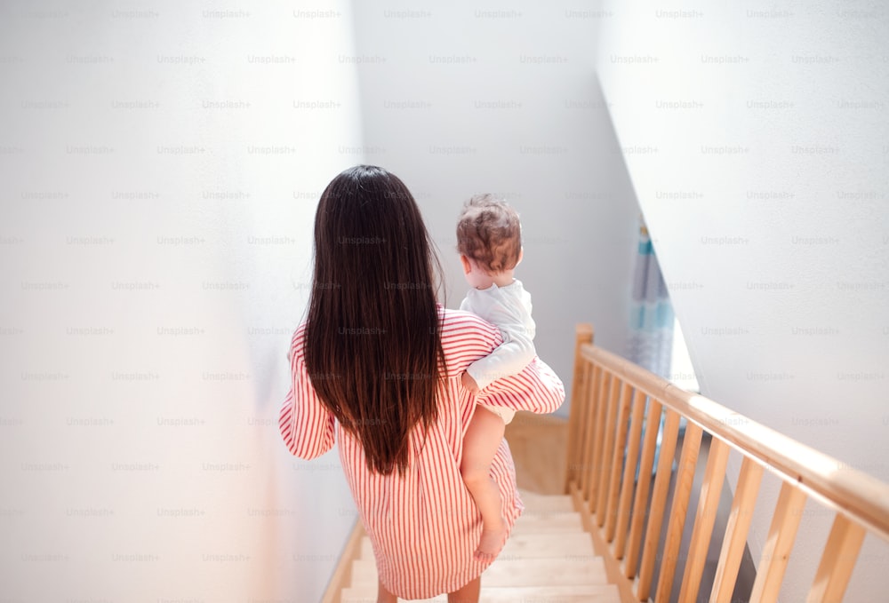 Rear view of young mother walking down the stairs indoors at home, carrying small toddler son.
