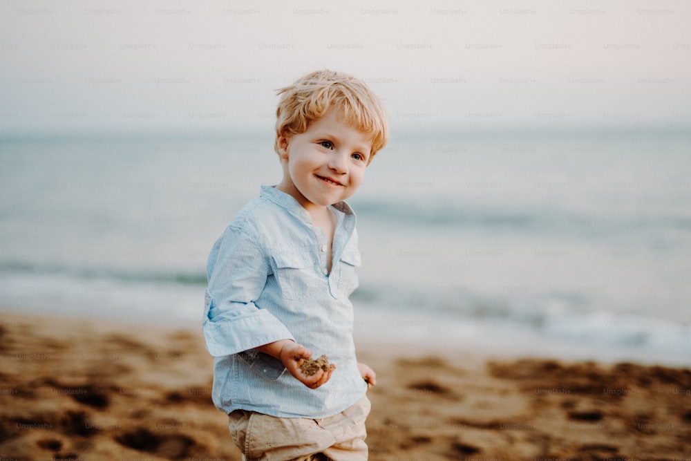 Um retrato de um menino pequeno alegre em pé na praia nas férias de verão.