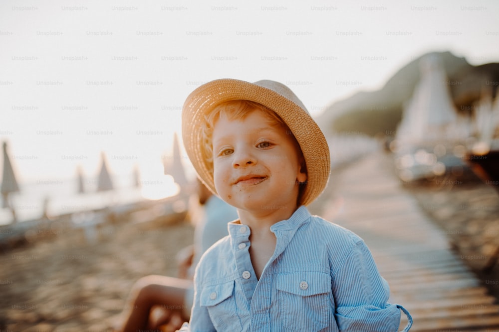 A small toddler boy with a hat standing on beach on summer holiday at sunset.