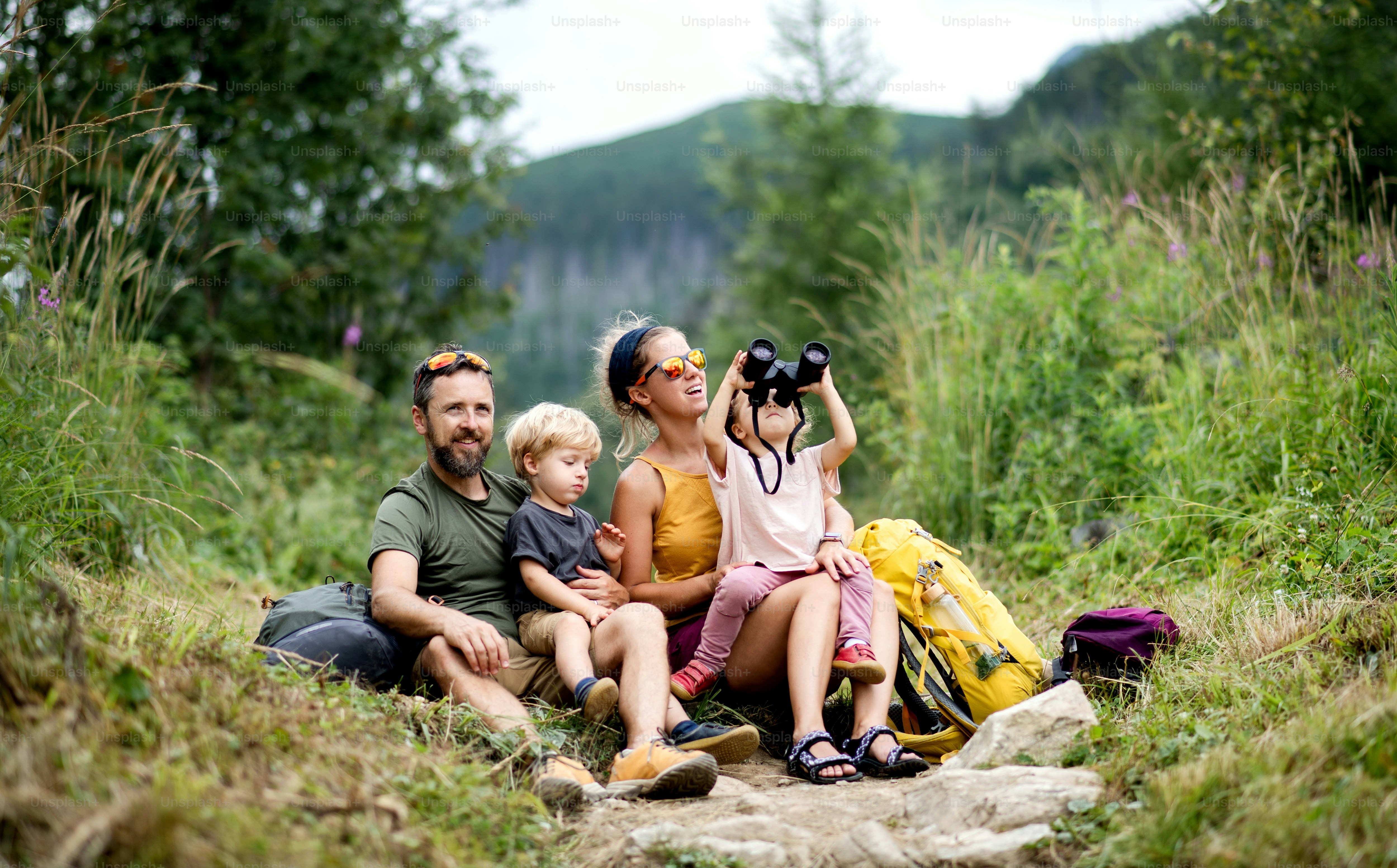 Front view of family with small children hiking outdoors in summer nature, sitting and resting