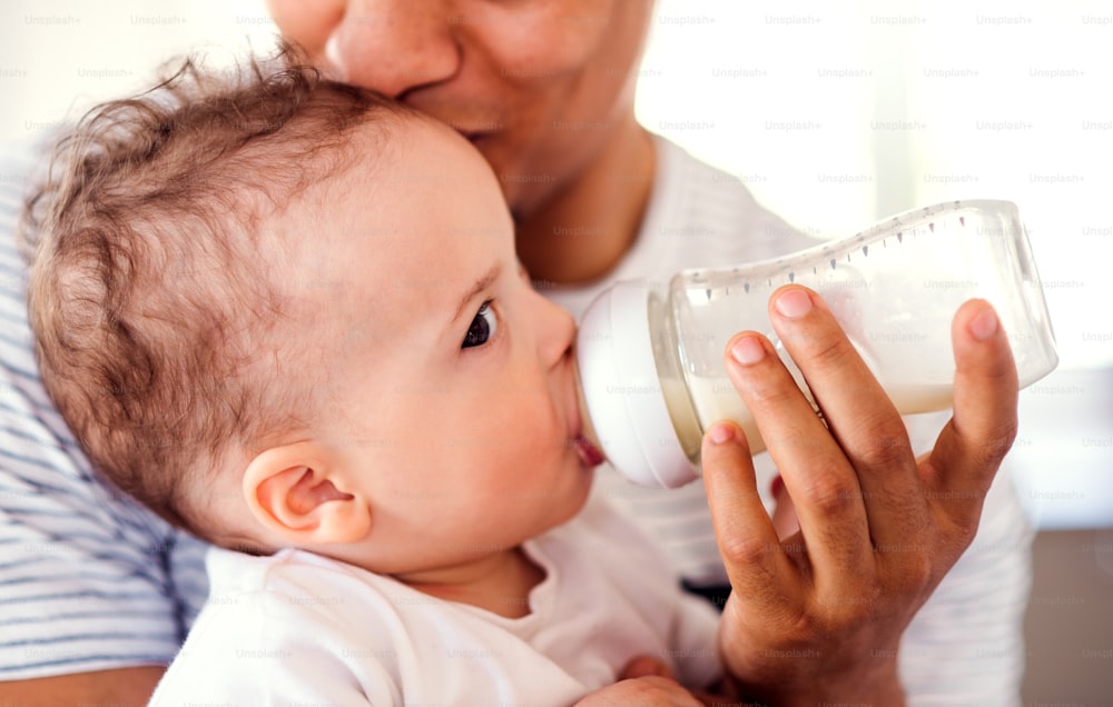 A midsection of unrecognizable father bottle feeding a small toddler son indoors at home.