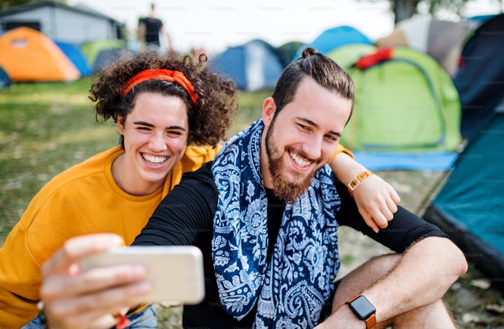Vista frontal do jovem casal no festival de verão ou férias de acampamento, tirando selfie com smartphone.