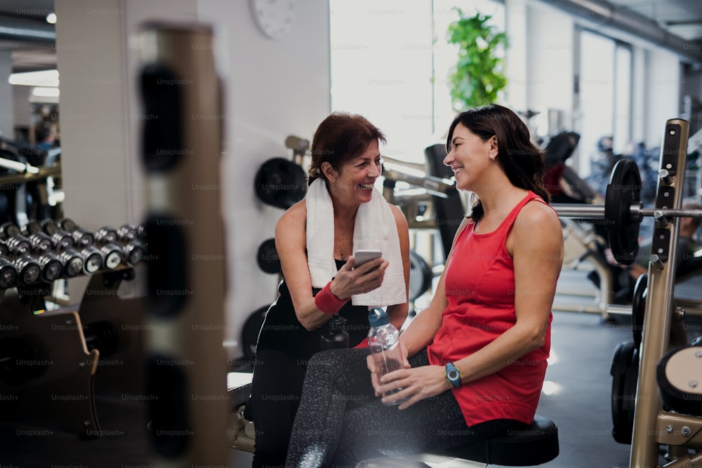 Two cheerful senior women in gym resting after doing exercise, holding smartphone and water bottle.