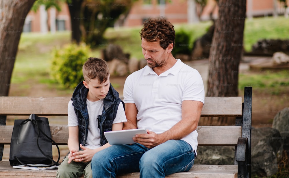 Father with son sitting on bench outdoors in park in town, using tablet.