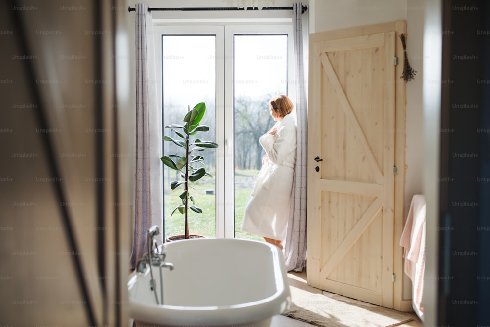 Una mujer joven con bata de baño de pie en el interior de un baño junto a una ventana por la mañana. Espacio de copia.
