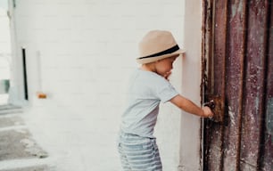 A small toddler boy with hat standing by a house in town on summer holiday, opening door. Copy space.