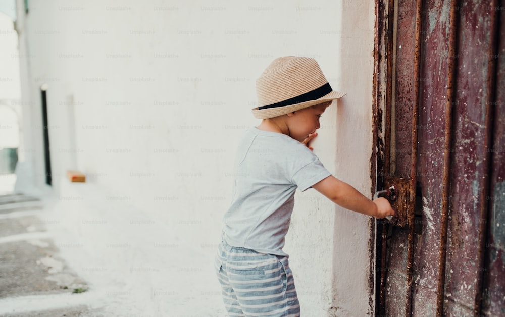 Un piccolo bambino con il cappello in piedi vicino a una casa in città durante le vacanze estive, aprendo la porta. Copia spazio.