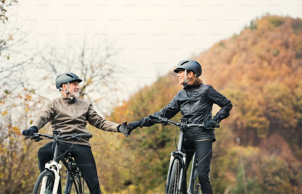 An active senior couple with helmets and electrobikes standing outdoors on a road in nature.