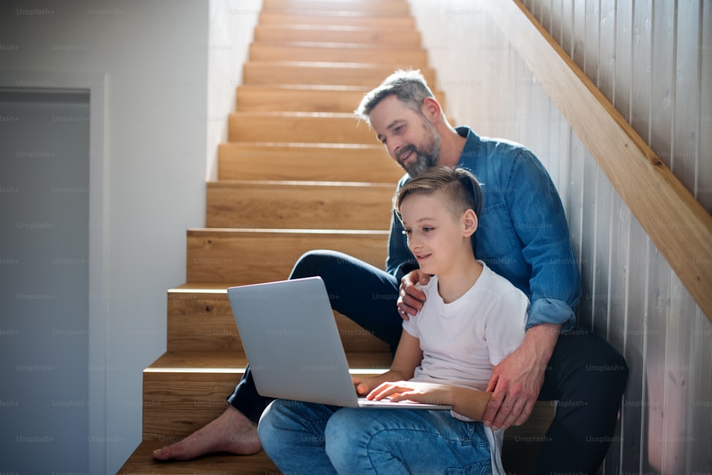 A mature father with small son sitting on the stairs indoors, using laptop.