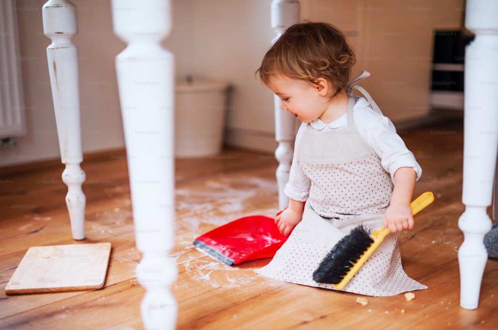 집에서 부엌에서 지저분한 바닥을 쓸고 있는 브러시와 쓰레받기를 가진 작은 유아 소녀.