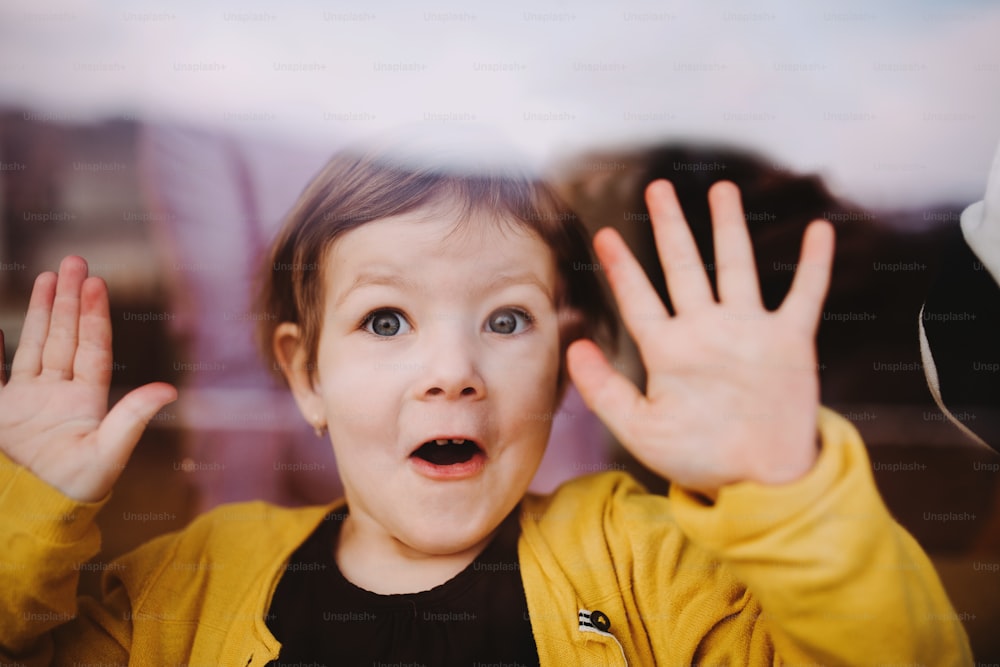 A close-up of a happy small girl looking through the window, shot through glass.