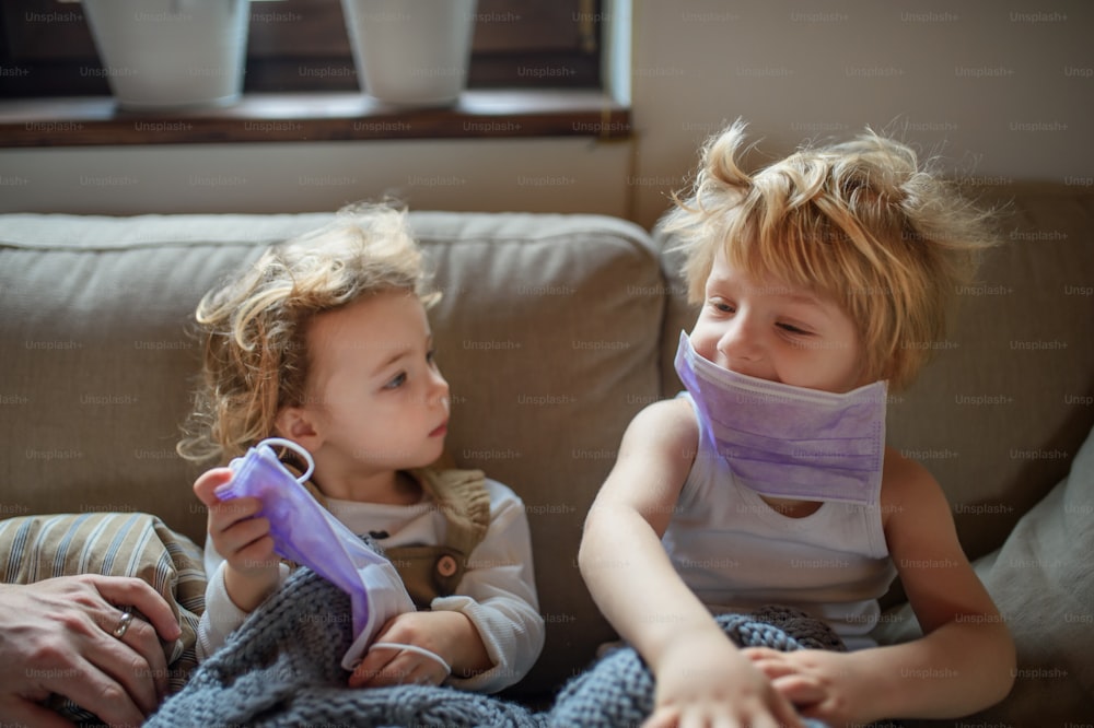 Front view of two small sick children with face mask at home sitting on sofa.