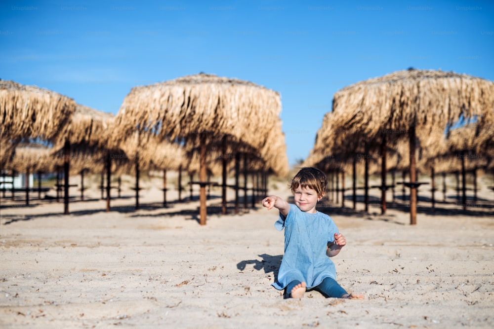Una bambina seduta sulla sabbia tra ombrelloni di paglia all'aperto sulla spiaggia.