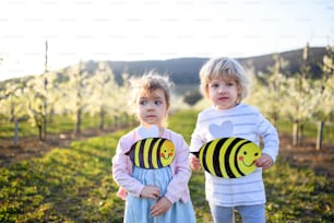 Front view of two small children standing outdoors in orchard in spring, with paper bees.