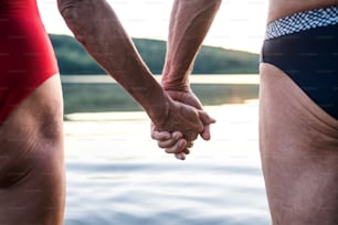 Midsection of senior couple in swimsuit by lake outdoors before swimming, holding hands.