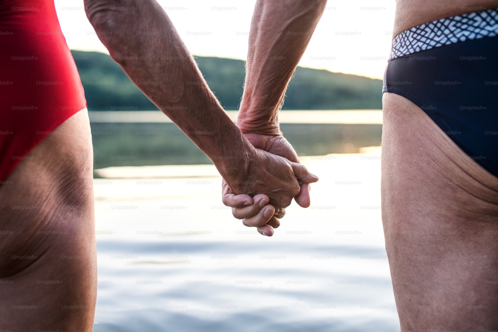 Midsection of senior couple in swimsuit by lake outdoors before swimming, holding hands.