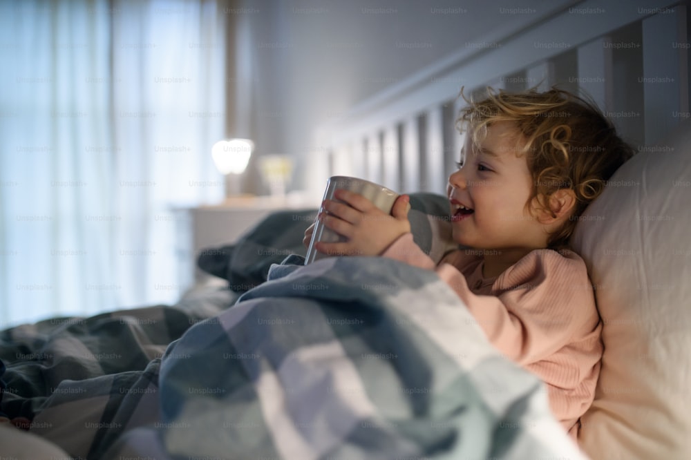 Small sick toddler girl lying in bed indoors at home, holding cup with tea.