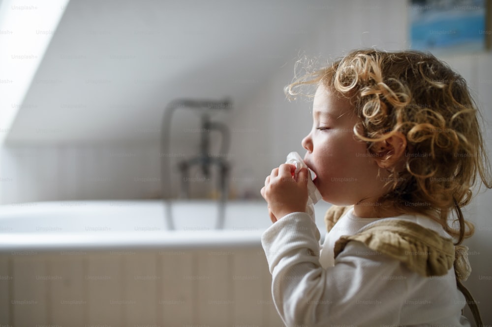 Vista lateral de una pequeña niña enferma con resfriado en casa sentada en el suelo, estornudando. Espacio de copia.