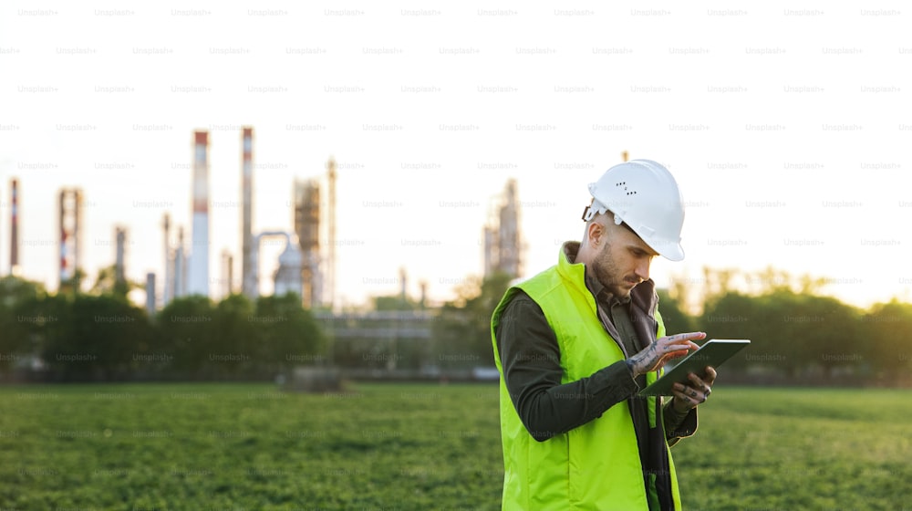 Serious young engineer with tablet standing outdoors by oil refinery. Copy space.