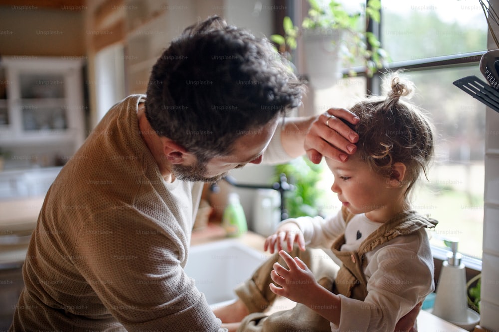 自宅の台所で病気の幼児の娘の額をチェックする父親。