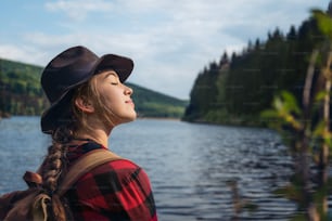 Mujer joven con los ojos cerrados de pie al aire libre junto al lago en un paseo por la naturaleza de verano, relajándose.