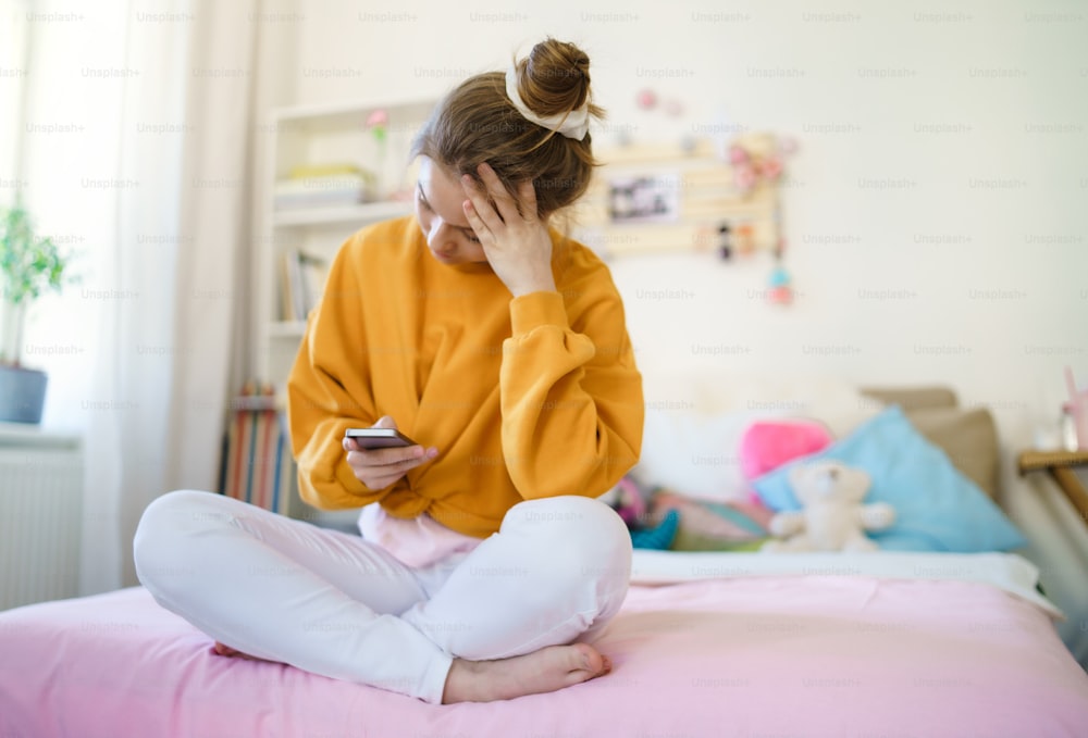 Sad young female student sitting on bed, using smartphone. Copy space.