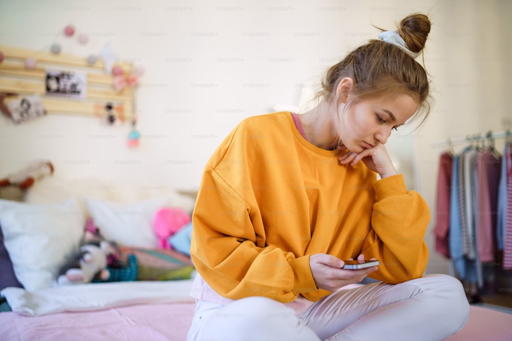 Triste giovane studentessa seduta sul letto, usando lo smartphone. Copia spazio.