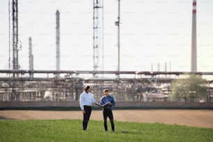 Dos jóvenes ingenieros parados al aire libre junto a una refinería de petróleo, discutiendo temas. Espacio de copia.