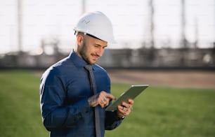 Young engineer with hard hat and tablet standing outdoors by oil refinery. Copy space.